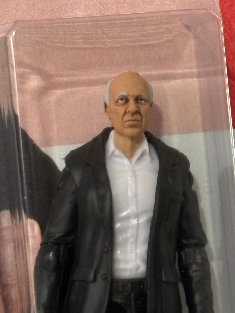 K&ouml;nnte beinhalten: Actionfigur eines &auml;lteren Mannes mit wei&szlig;em Haar, bekleidet mit einer schwarzen Jacke &uuml;ber einem wei&szlig;en Hemd mit Kragen. Die Figur befindet sich in einer transparenten Kunststoffverpackung mit rosa Hintergrund.
