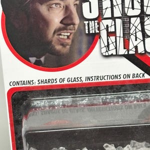 K&ouml;nnte beinhalten: Eine Produktverpackungskarte mit dem Portr&auml;t eines Mannes und dem Text SHOOT THE GLASS. Die Verpackung enth&auml;lt eine durchsichtige Kunststoffblisterpackung mit kleinen, klaren, gezackten Glassplittern. Der Text CONTAINS: SHARDS OF GLASS, INSTRUCTIONS ON BACK ist in Schwarz auf der wei&szlig;en Kartenoberfl&auml;che aufgedruckt.