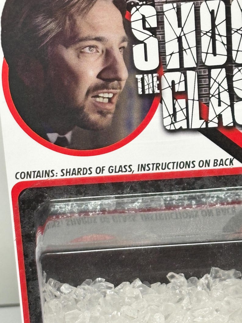 K&ouml;nnte beinhalten: Eine Produktverpackungskarte mit dem Portr&auml;t eines Mannes und dem Text SHOOT THE GLASS. Die Verpackung enth&auml;lt eine durchsichtige Kunststoffblisterpackung mit kleinen, klaren, gezackten Glassplittern. Der Text CONTAINS: SHARDS OF GLASS, INSTRUCTIONS ON BACK ist in Schwarz auf der wei&szlig;en Kartenoberfl&auml;che aufgedruckt.