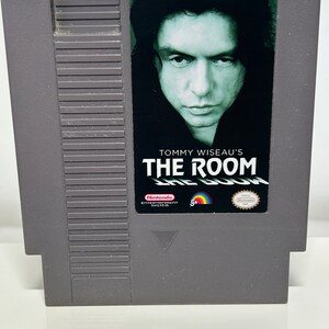 K&ouml;nnte beinhalten: Eine graue Nintendo Entertainment System-Spielkassette mit einem schwarzen Etikett. Das Etikett zeigt ein Foto von Tommy Wiseau, den Text "Tommy Wiseau's The Room" und das Nintendo Entertainment System-Logo.