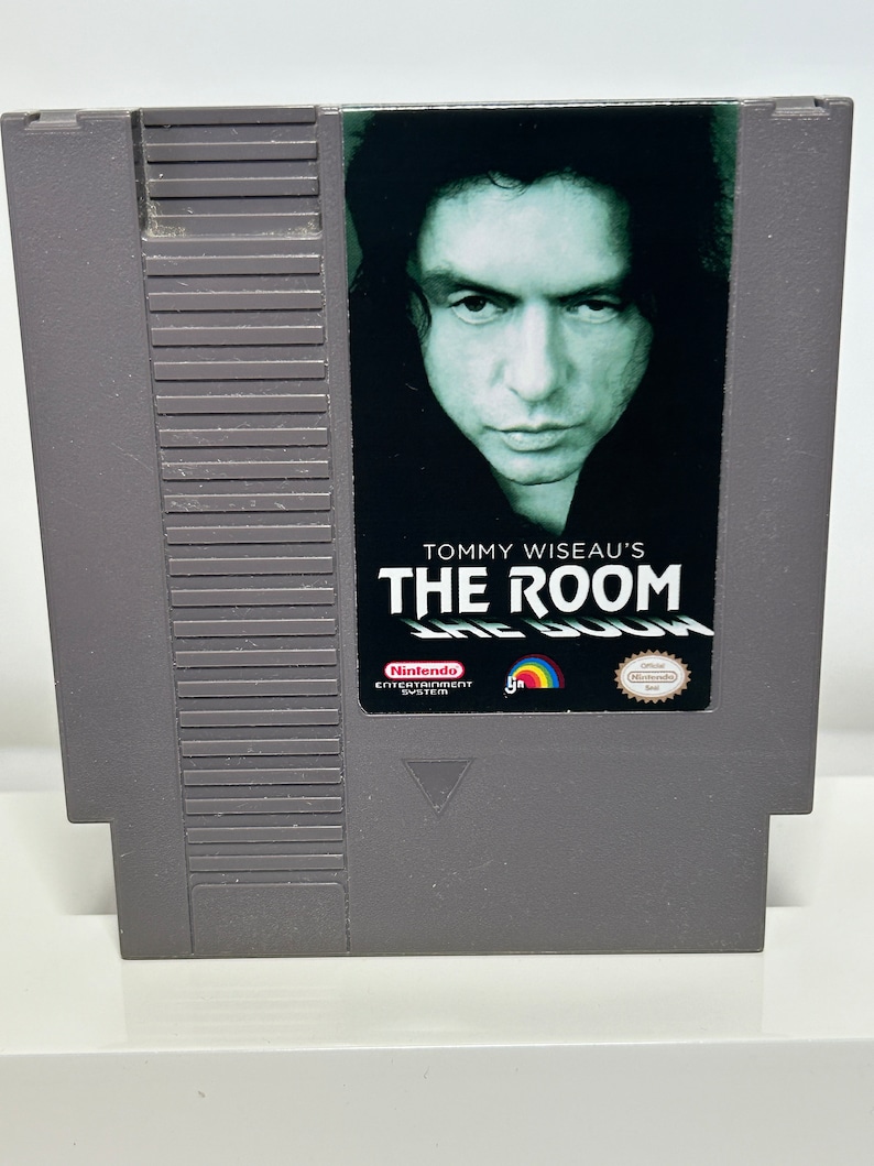 K&ouml;nnte beinhalten: Eine graue Nintendo Entertainment System-Spielkassette mit einem schwarzen Etikett. Das Etikett zeigt ein Foto von Tommy Wiseau, den Text "Tommy Wiseau's The Room" und das Nintendo Entertainment System-Logo.