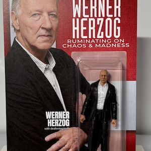K&ouml;nnte beinhalten: Sammelfigur von Werner Herzog in einer transparenten Kunststoffverpackung. Die Verpackung zeigt ein Foto von Herzog und den Text "WERNER HERZOG RUMINATING ON CHAOS & MADNESS". Die Figur tr&auml;gt einen schwarzen Mantel und eine schwarze Hose.