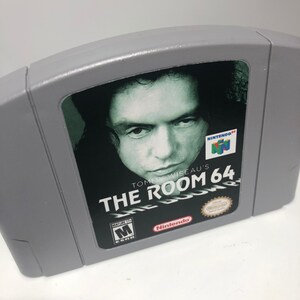 K&ouml;nnte beinhalten: Eine graue Nintendo 64-Spielkassette mit einem Schwarzwei&szlig;bild von Tommy Wiseau auf der Vorderseite. Der Spieltitel lautet "Tommy Wiseau's The Room 64" und das Spiel ist von der ESRB mit "M" f&uuml;r Mature bewertet.