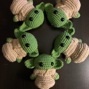 K&ouml;nnte beinhalten: F&uuml;nf geh&auml;kelte Pl&uuml;schtiere in Form einer beliebten fiktiven Figur. Die Spielzeuge sind gr&uuml;n und beige und haben gro&szlig;e schwarze Augen. Die Spielzeuge sind in einem Kreis angeordnet.