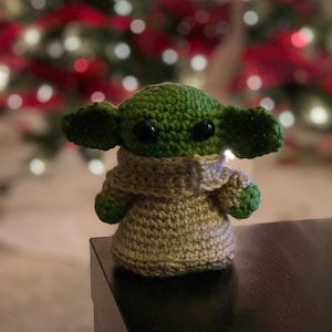 K&ouml;nnte beinhalten: Eine geh&auml;kelte Yoda-Puppe in Gr&uuml;n und Braun, die auf einem schwarzen Tisch vor einem Weihnachtsbaum mit roten und wei&szlig;en Kugeln sitzt.