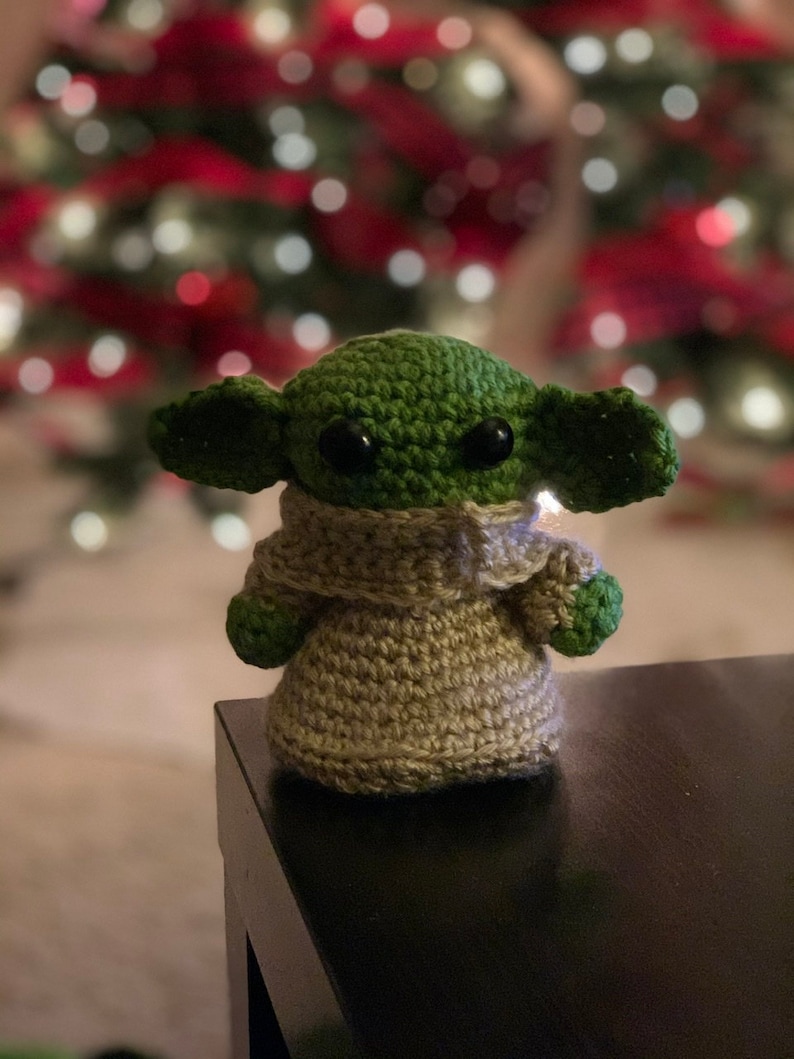 K&ouml;nnte beinhalten: Eine geh&auml;kelte Yoda-Puppe in Gr&uuml;n und Braun, die auf einem schwarzen Tisch vor einem Weihnachtsbaum mit roten und wei&szlig;en Kugeln sitzt.