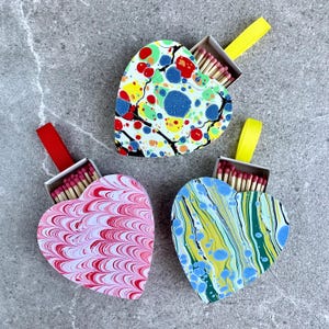 May include: Three heart-shaped matchboxes with colorful marbled designs. Each box has a ribbon attached and contains matches.