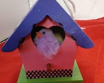 Könnte beinhalten: Ein dekoratives Vogelhaus mit einem rosa Körper, einem blauen Dach und einer grünen Basis. Das Vogelhaus hat eine herzförmige Öffnung mit einem flauschigen lila Vogel im Inneren. Eine weiße Schnur ist zum Aufhängen befestigt.