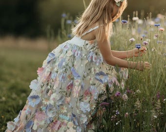 Può includere: Un abito senza maniche con uno strato superiore trasparente decorato con numerosi fiori in tessuto tridimensionali nelle tonalità del rosa, blu e bianco. L'abito presenta un corpetto aderente e una gonna ampia e fluida fino a terra.