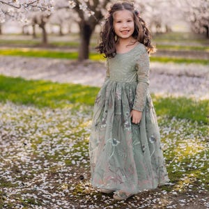 May include: A young girl in a sage green dress with floral embroidery stands in a field of white flower petals. The dress has long sleeves and a flowing skirt. The background features blossoming trees and a green lawn.