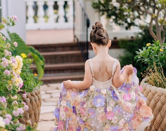 May include: A girl in a floral dress walks along a stone path. The dress is adorned with pastel-coloured flowers in shades of pink, purple, and peach. The dress has a cream-coloured bodice and straps. The background includes greenery and a fountain.