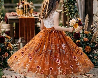 Può includere: Un abito lungo fino ai piedi con un corpetto in pizzo bianco e un'ampia gonna in tulle color arancio bruciato, decorata con intricati ricami floreali multicolori. L'abito include un nastro arancione abbinato annodato in vita.
