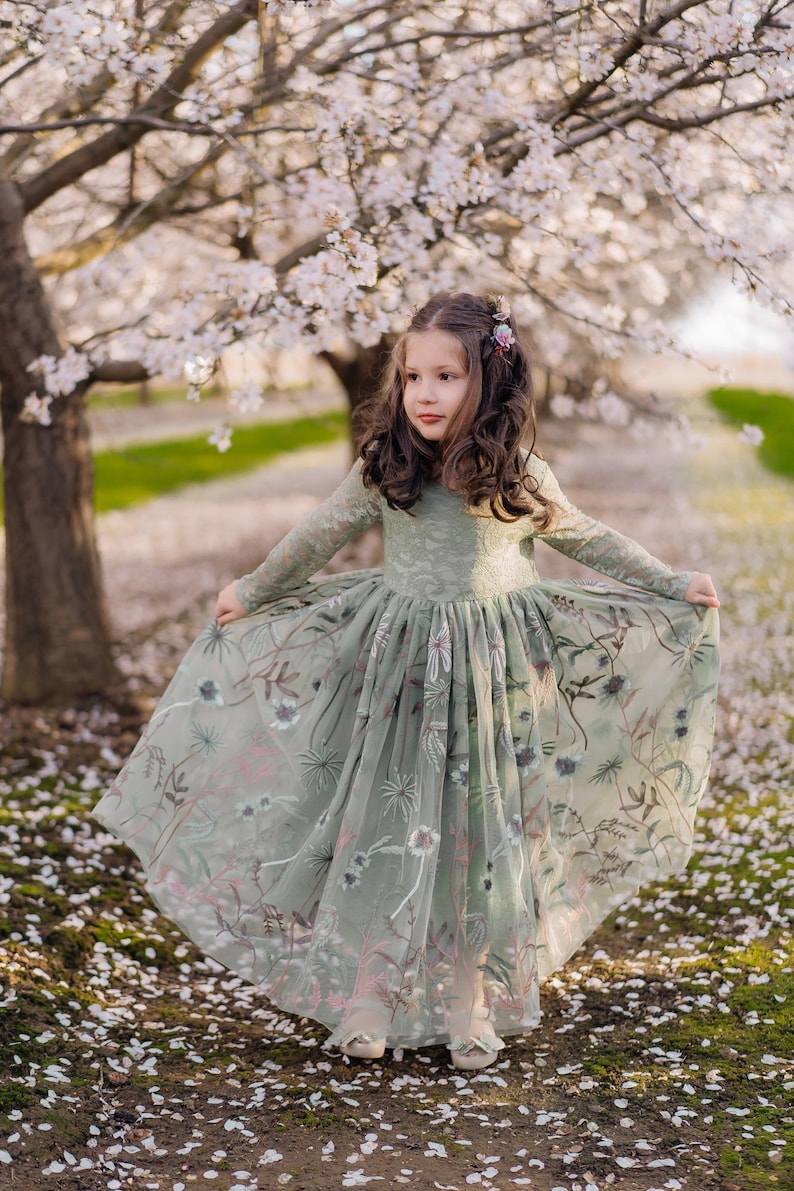 Pu&ograve; includere: Un abito a maniche lunghe color verde salvia con corpetto in pizzo e gonna in tulle fluida decorata con intricati ricami floreali nei toni del rosa, bianco e verde scuro. L'abito presenta uno scollo rotondo e una silhouette ampia.
