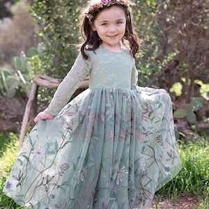 Pu&ograve; includere: Un abito a maniche lunghe color verde salvia con corpetto in pizzo e gonna in tulle caratterizzata da intricati ricami floreali nei toni del marrone, rosa e bianco. L'abito &egrave; abbinato a una corona di piccoli fiori viola, rosa e bianchi.