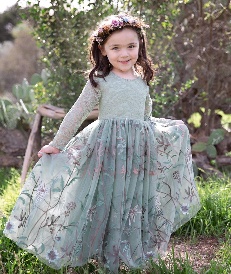 Pu&ograve; includere: Un abito a maniche lunghe color verde salvia con corpetto in pizzo e gonna in tulle caratterizzata da intricati ricami floreali nei toni del marrone, rosa e bianco. L'abito &egrave; abbinato a una corona di piccoli fiori viola, rosa e bianchi.