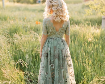 May include: A sage green dress with floral embroidery, featuring a lace bodice and a flowing tulle skirt. The dress has short sleeves and a V-neck back. The image is taken outdoors in a field of tall grass.