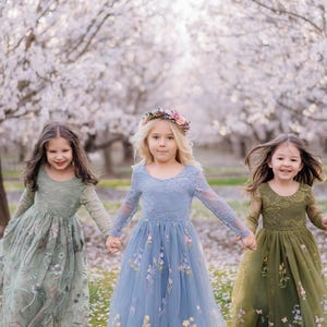 Pu&ograve; includere: Tre abiti a maniche lunghe in verde salvia, blu polvere e verde oliva, ciascuno con scollo rotondo, dettagli in pizzo sul corpetto e gonna in tulle decorata con delicati ricami floreali, indossati da tre bambini in un frutteto.