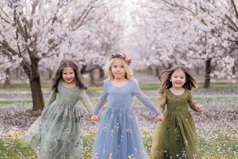 Pu&ograve; includere: Tre abiti a maniche lunghe in verde salvia, blu polvere e verde oliva, ciascuno con scollo rotondo, dettagli in pizzo sul corpetto e gonna in tulle decorata con delicati ricami floreali, indossati da tre bambini in un frutteto.