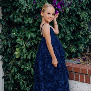 May include: A young girl in a navy blue floral-appliqué dress with spaghetti straps. The dress has a full skirt and is knee-length. She is standing in front of a green bush with purple flowers. The girl has blonde hair and is wearing a bracelet.