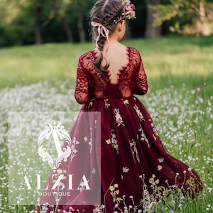 Könnte beinhalten: Ein junges Mädchen trägt ein burgunderfarbenes Tüllkleid mit Blumenstickerei und Spitzenärmeln. Sie steht auf einem Feld mit weißen Blumen und trägt eine Blumenkrone im Haar.