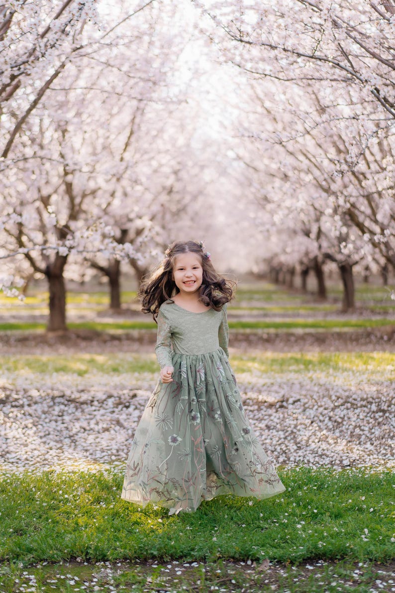 Pu&ograve; includere: Un abito a maniche lunghe color verde salvia con corpetto aderente e gonna in tulle fluida decorata con intricati ricami floreali nei toni del bianco, marrone e verde tenue, che arriva fino alle caviglie con scollo rotondo.
