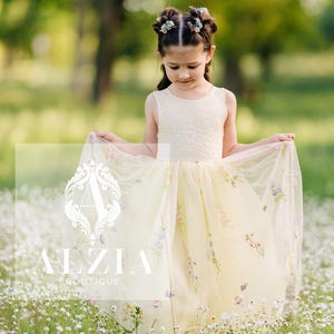 May include: A young girl wearing a yellow tulle dress with floral embroidery and a white lace bodice. She is standing in a field of white flowers.