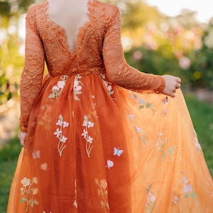 Puede incluir: Un vestido de manga larga en tono naranja quemado con un corpiño de encaje y escote en V en la espalda. La falda está hecha de tul superpuesto y está adornada con patrones florales bordados y pequeños motivos de mariposas en tonos suaves.