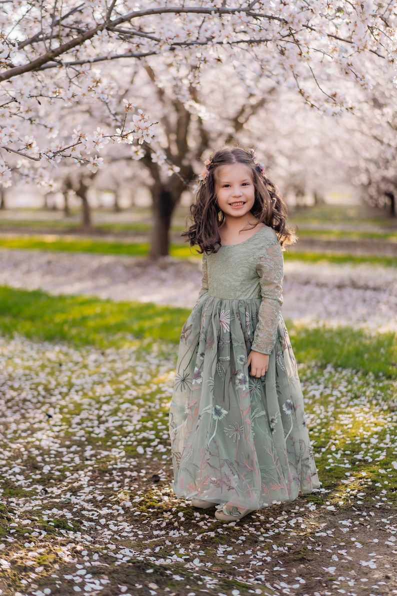 May include: A young girl in a sage green dress with floral embroidery stands in a field of white flower petals. The dress has long sleeves and a flowing skirt. The background features blossoming trees and a green lawn.