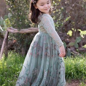 Pu&ograve; includere: Un abito a maniche lunghe color verde salvia con intricati dettagli in pizzo sul corpetto e sulle maniche, abbinato a una gonna in tulle fluida decorata con motivi floreali e di viti ricamati in tonalit&agrave; marrone, rosa e verde, e una corona di fiori.