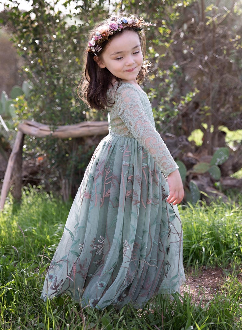 Pu&ograve; includere: Un abito a maniche lunghe color verde salvia con intricati dettagli in pizzo sul corpetto e sulle maniche, abbinato a una gonna in tulle fluida decorata con motivi floreali e di viti ricamati in tonalit&agrave; marrone, rosa e verde, e una corona di fiori.