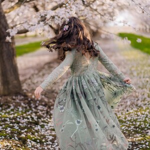 Pu&ograve; includere: Un abito verde salvia a maniche lunghe con corpetto in pizzo e gonna in tulle fluida decorata con intricati ricami floreali nei toni del rosa, bianco e verde scuro, con scollo a V sulla schiena e una silhouette ampia e stratificata.