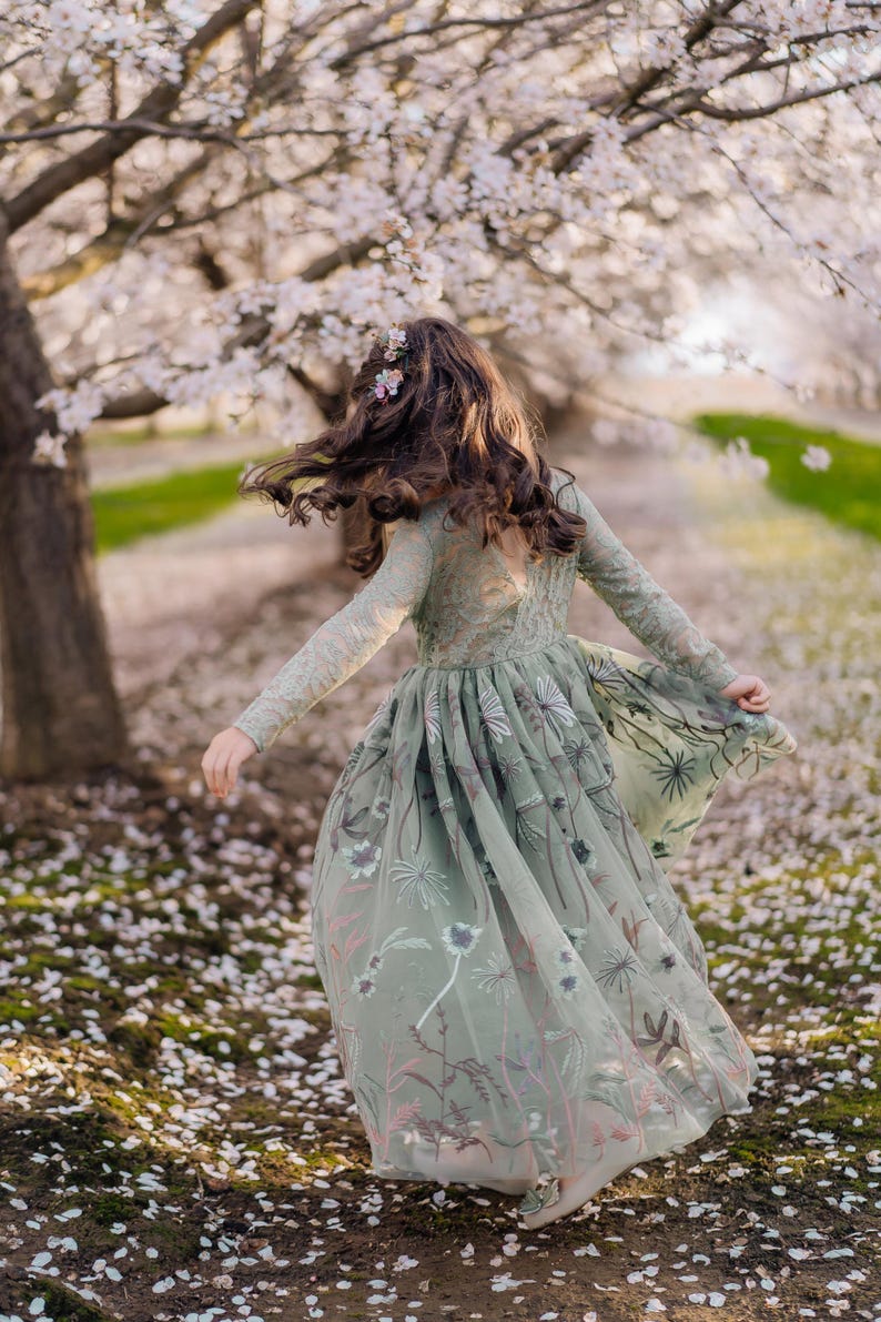 Pu&ograve; includere: Un abito verde salvia a maniche lunghe con corpetto in pizzo e gonna in tulle fluida decorata con intricati ricami floreali nei toni del rosa, bianco e verde scuro, con scollo a V sulla schiena e una silhouette ampia e stratificata.