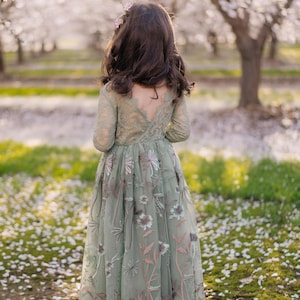 Pu&ograve; includere: Un abito lungo color verde salvia con maniche lunghe in pizzo, scollo a V sulla schiena e uno strato di tulle trasparente ricamato con delicati motivi floreali nei toni del rosa, bianco e verde, indossato da un bambino dai capelli scuri ondulati.