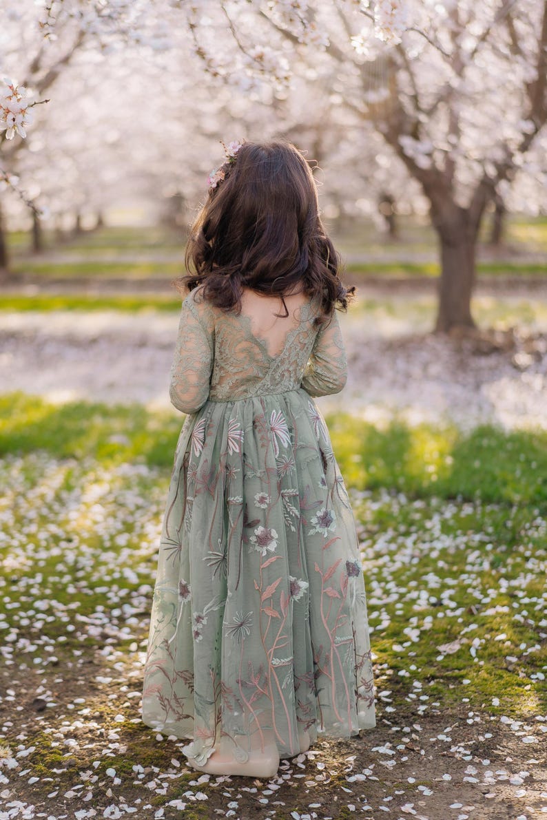 Pu&ograve; includere: Un abito lungo color verde salvia con maniche lunghe in pizzo, scollo a V sulla schiena e uno strato di tulle trasparente ricamato con delicati motivi floreali nei toni del rosa, bianco e verde, indossato da un bambino dai capelli scuri ondulati.