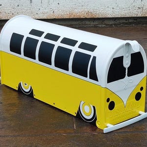 Puede incluir: Un buzón amarillo y blanco con forma de autobús vintage. El autobús tiene ventanas y ruedas negras, con un parachoques delantero blanco. El buzón está diseñado para parecerse a un autobús escolar clásico.