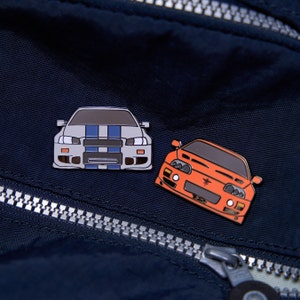 以下が含まれることがあります： 2つの車の形をしたエナメルピン。1つは青いストライプのシルバー、もう1つはオレンジ色です。どちらのピンも黒いアウトラインで、濃いネイビーブルーの背景に配置されています。ピンはヘッドライトやその他の車の特徴で詳細に描かれています。