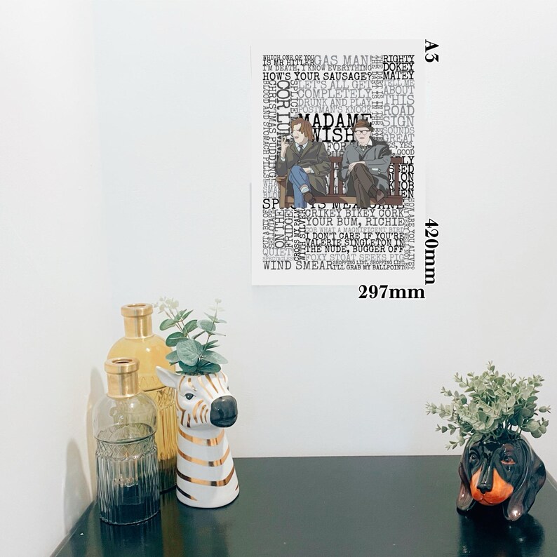 K&ouml;nnte beinhalten: Ein gerahmter A3-Druck (420 mm x 297 mm) mit zwei Figuren auf einer Bank, den Worten "MADAME WISH" und anderen Phrasen in einer Collage. Dekorative Elemente sind eine zebra-gestreifte Vase, Glasflaschen und ein Dackel-Pflanzgef&auml;&szlig;.
