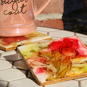 May include: Set of four square coasters featuring abstract artwork in shades of yellow, red, and brown. The coasters have a gold trim and are made of ceramic.