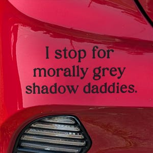 Puede incluir: Parachoques rojo de coche con el texto "I stop for morally grey shadow daddies" en letras negras. El parachoques tiene una rejilla negra y un embellecedor negro.