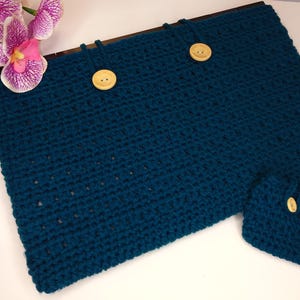 May include: Teal crocheted laptop sleeve with two wooden buttons. A matching small accessory, also teal and crocheted, is included. The sleeve is rectangular and has a wooden bar at the top. The background is white with pink and purple flowers.