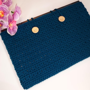 Può includere: Una custodia per laptop all'uncinetto blu teal con due bottoni di legno. La custodia ha una cornice in legno scuro ed è decorata con un rametto di orchidee rosa e viola.