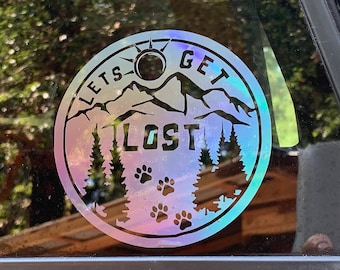 May include: A round, iridescent sticker featuring a mountain scene with the text "Let's Get Lost". The sticker also includes a sun, trees, and paw prints.