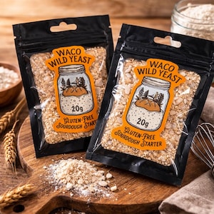 Könnte beinhalten: Zwei schwarze Beutel mit glutenfreiem Sauerteigstarter von Waco Wild Yeast, jeweils 20g. Die Etiketten zeigen eine Gläser-Illustration mit einem Brotlaib. Die Beutel liegen auf einem Holzschneidebrett mit verstreutem Starter.