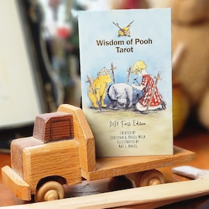 Puede incluir: Un camión de juguete de madera transporta una baraja de cartas "Wisdom of Pooh Tarot". La baraja presenta ilustraciones de los personajes de Winnie the Pooh. El camión es de madera marrón clara y oscura. Es una primera edición de 2023.