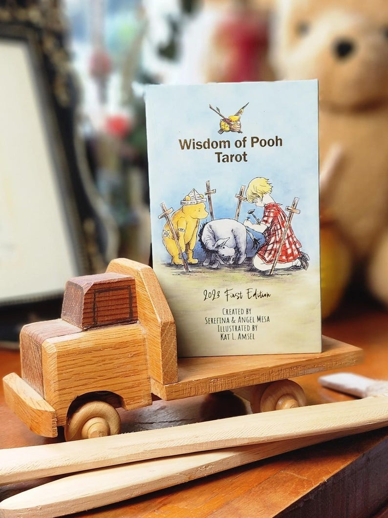 May include: A rectangular Wisdom of Pooh Tarot deck box featuring a light blue background with an illustration of Winnie the Pooh, Eeyore, and Christopher Robin. The text reads Wisdom of Pooh Tarot, 2023 First Edition, Serefina & Angel Mesa, Kat L. Amsel.
