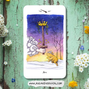 May include: A rectangular tarot card featuring a watercolor illustration of a hand emerging from clouds to hold a sword topped with a golden crown and green leaves. A small yellow bear wearing a paper hat stands below. The text reads Ace.