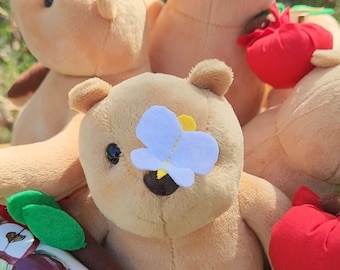 May include: A collection of plush teddy bears in various poses. Each bear is light brown with a flower or butterfly accent. Some bears hold red apple-shaped objects. The bears are arranged in a woven basket, creating a whimsical and charming display.