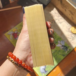 May include: A rectangular deck of cards featuring metallic gold gilded edges. The cards are stacked neatly together, showcasing the reflective gold finish on the side of the deck. The deck is held firmly in place, displaying the uniform thickness and smooth, shiny surface of the gilded card edges.