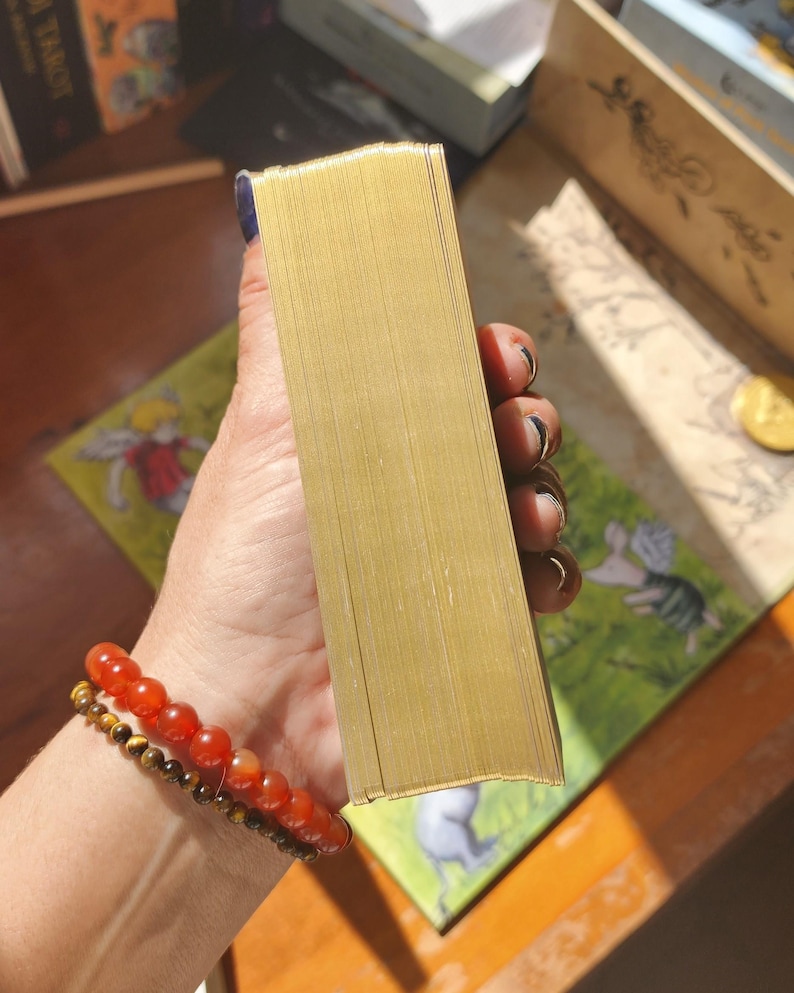 May include: A rectangular deck of cards featuring metallic gold gilded edges. The cards are stacked neatly together, showcasing the reflective gold finish on the side of the deck. The deck is held firmly in place, displaying the uniform thickness and smooth, shiny surface of the gilded card edges.