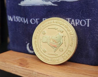 May include: A gold-colored coin with a Winnie the Pooh design, featuring text that reads "A day without a drop of honey" and "2023". The coin is set against a wooden surface and a blue background with the words "Wisdom of Tarot".